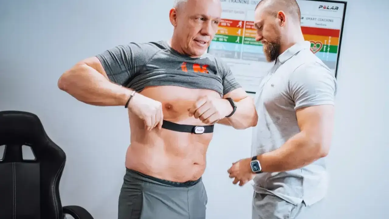 Mann legt Brustgurt für Muskelerhalt Training an, Trainer hilft.