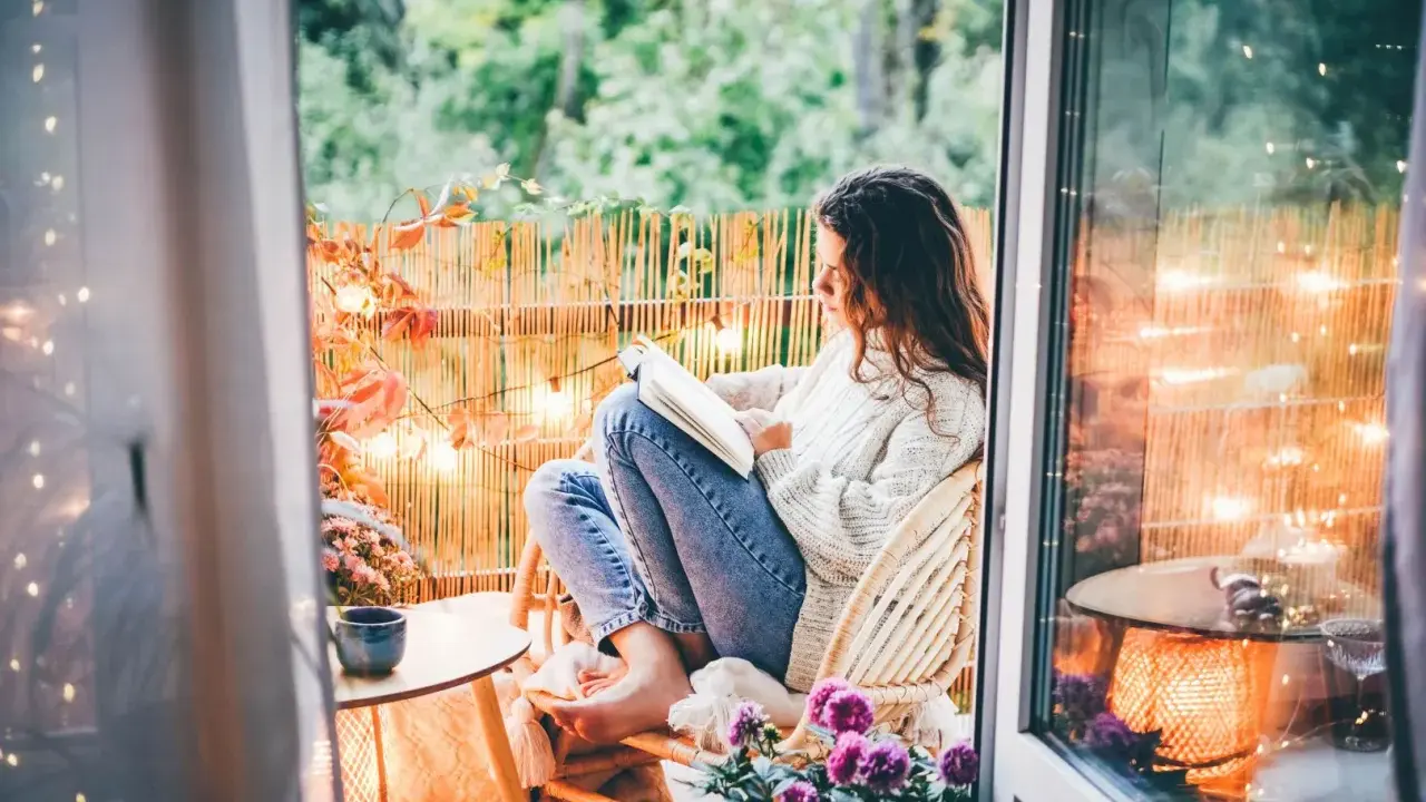 Przytulny jesienny balkon. Dziewczyna w swetrze czyta książkę, otoczona jesiennymi liśćmi i ciepłym światłem lampek.