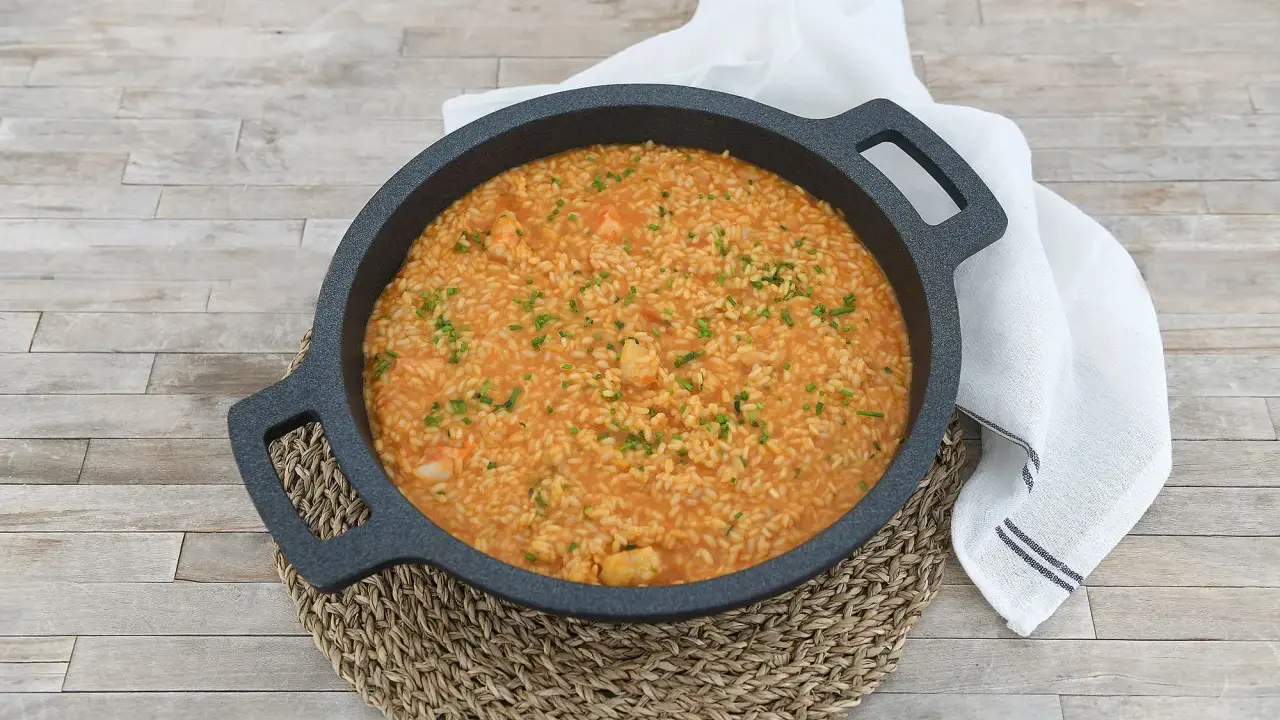 Arroz cremoso con gambones, adornado con cebollino fresco, servido en una paellera sobre una mesa de madera.