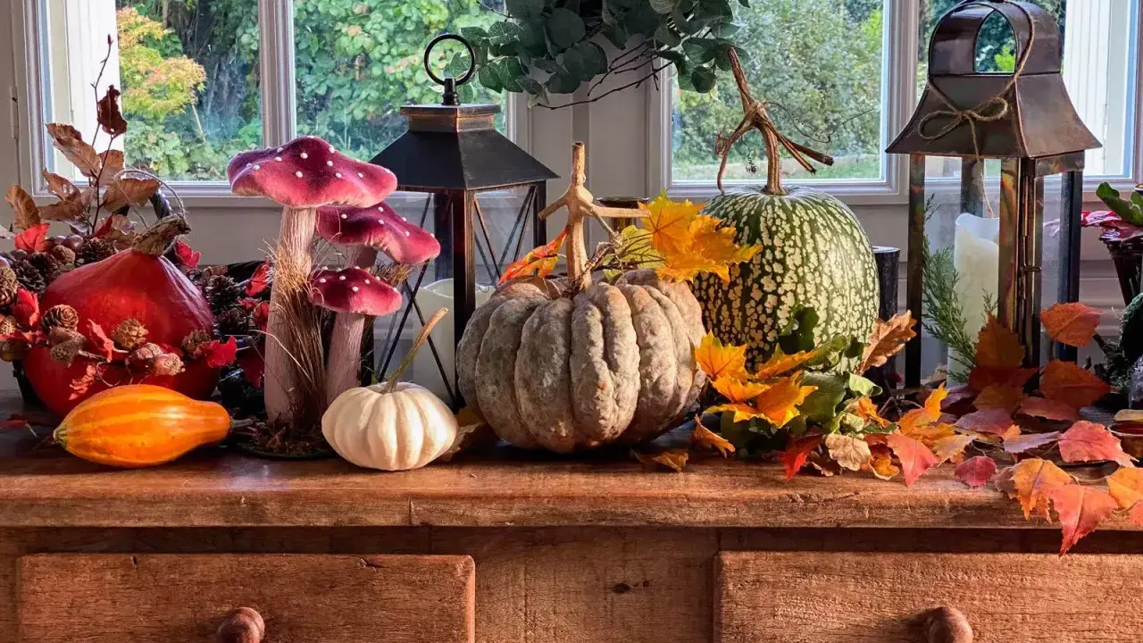 Décoration automne facile à faire : citrouilles, lanternes, fausses champignons et feuilles d'automne sur un buffet en bois devant une fenêtre.