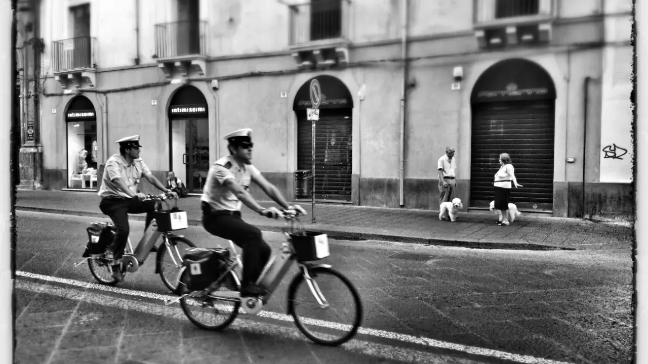 Due agenti in bicicletta pattugliano una strada italiana. Sul marciapiede, una coppia passeggia con i loro cani. Street photography Italia.