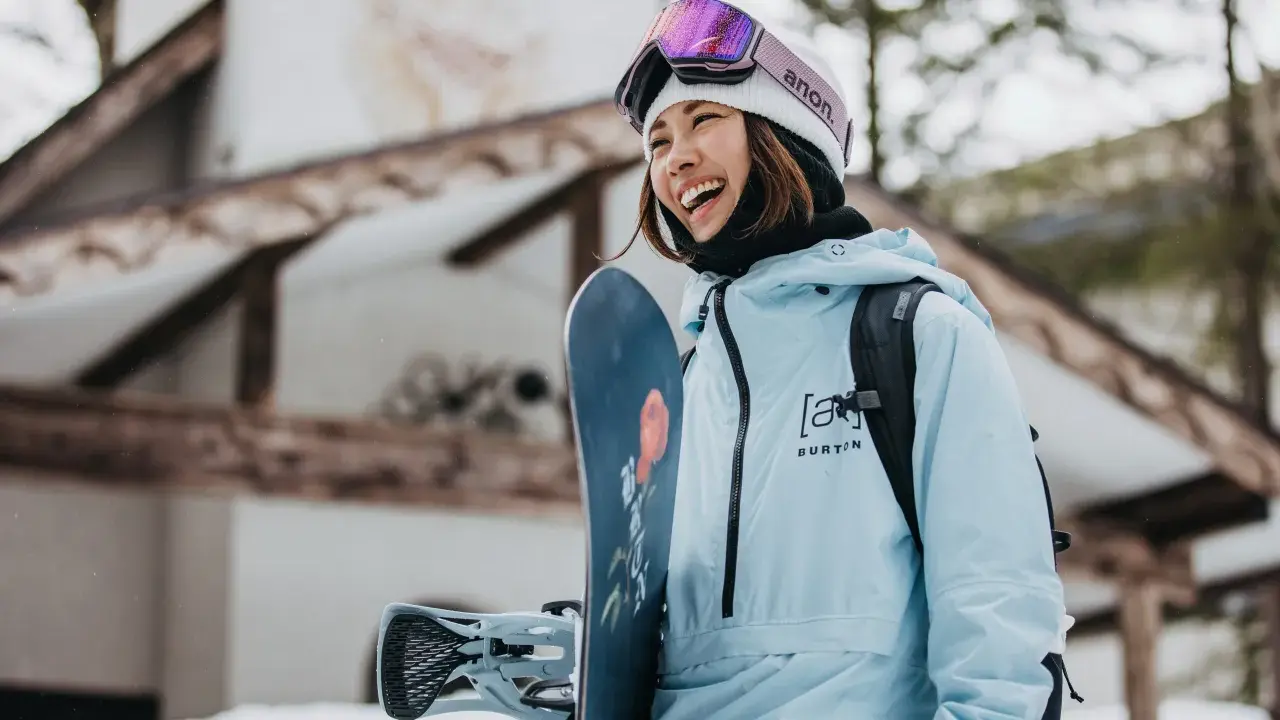 Uśmiechnięta snowboardzistka w jasnoniebieskim anoraku i goglach Anon. Gotowa na stok, trzyma deskę Burton.