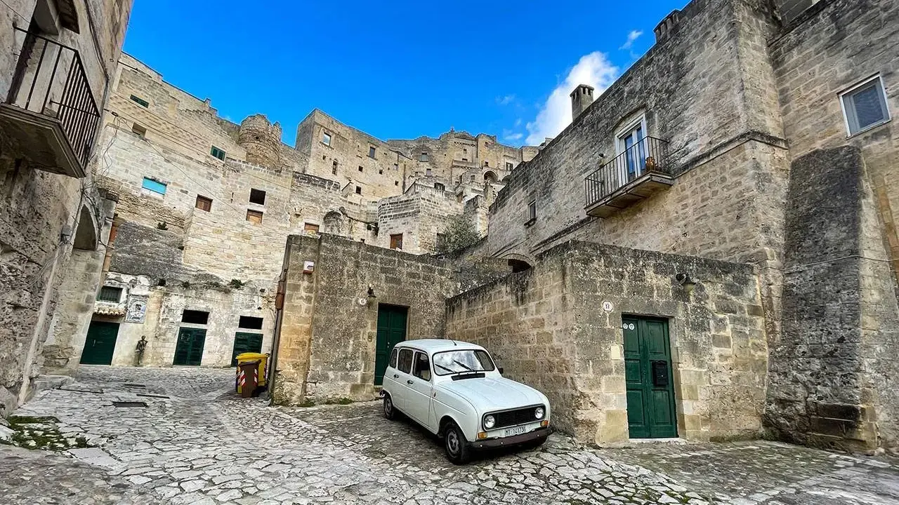 Un'auto d'epoca bianca parcheggiata tra le antiche case scavate nella roccia dei Sassi di Matera, sotto un cielo azzurro.