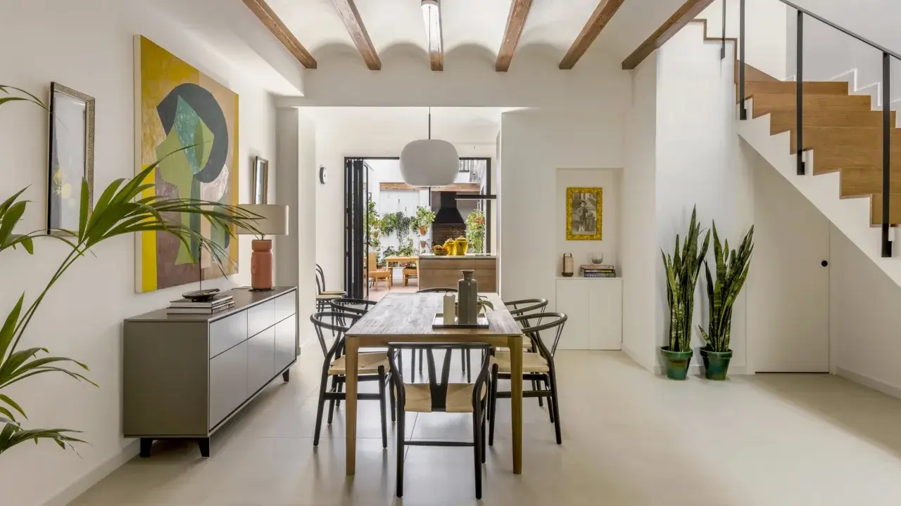 Comedor moderno en una casa entre medianeras, con vigas de madera en el techo, mesa de madera, sillas negras y una escalera de madera.