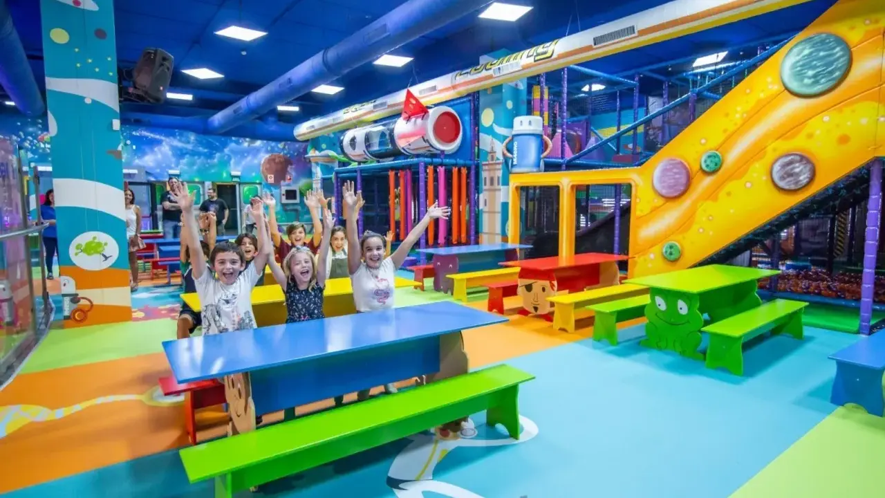 Un grupo de niños alegres levanta las manos en un vibrante centro comercial con juegos para niños, con toboganes, túneles y coloridos muebles.