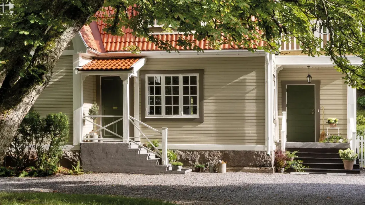 Ett charmigt hus med tegelrött tak och ljusgul fasad, redo att måla om huset. En stor ek skuggar entrén.
