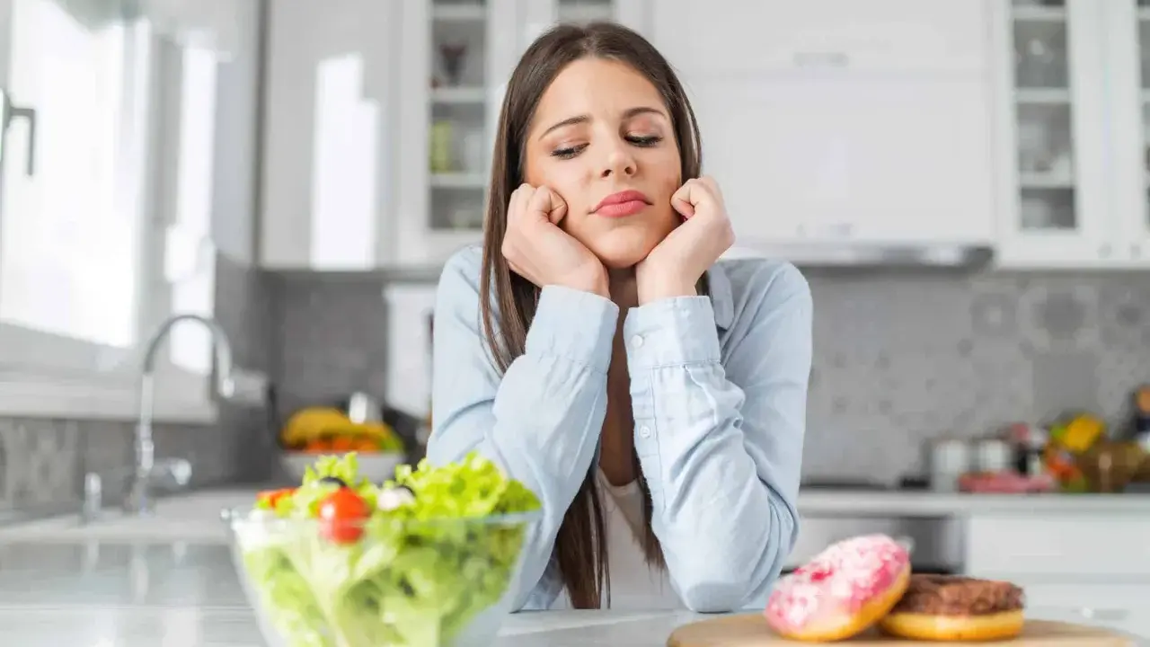Femme triste face à une salade et des donuts, pensant aux aliments à éviter pour la dermite séborrhéique.