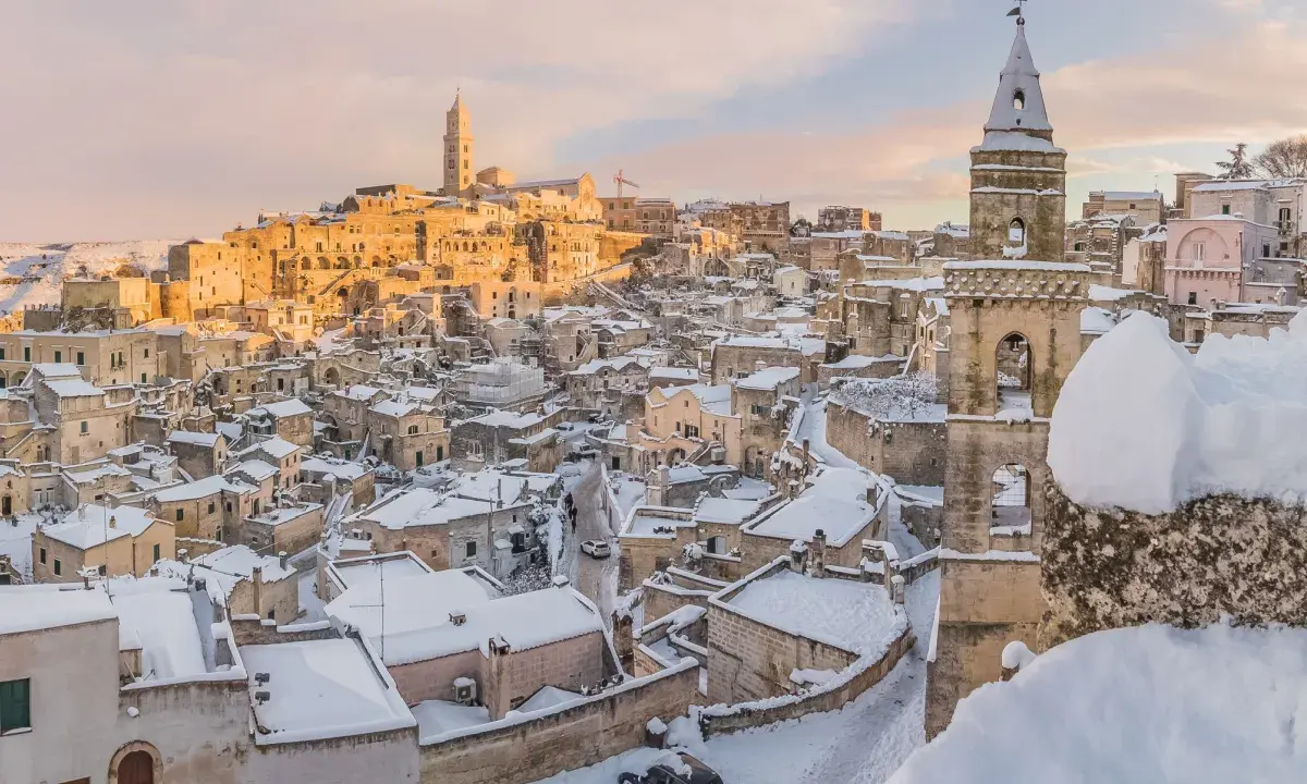 Matera innevato, un incanto tra i **posti da visitare in inverno Italia**. Case antiche e chiese sotto la neve, con il sole che illumina il paesaggio.