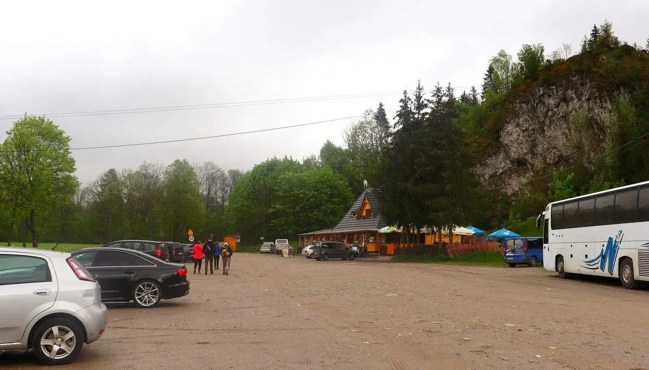 Parking przy Wąwozie Homole. Samochody, autokar i budynek zadaszony, otoczony drzewami, u podnóża skalnego zbocza.