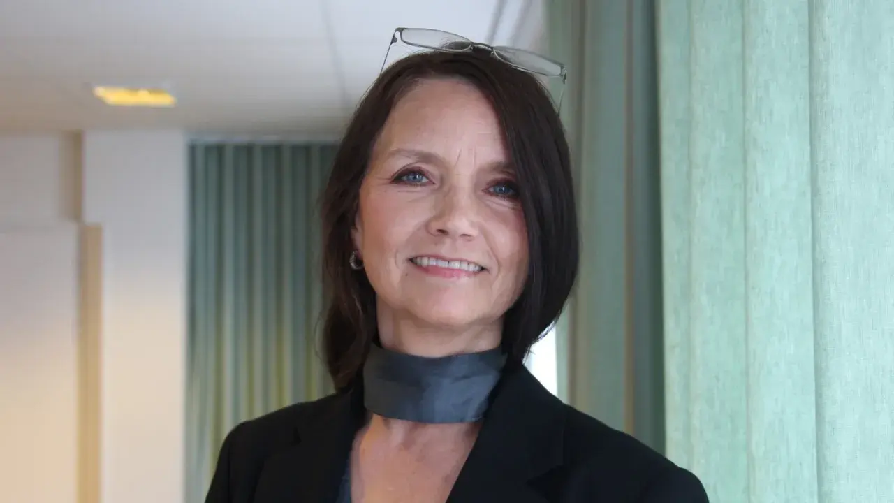 En kvinna med glasögon på huvudet ler. Hon bär en svart kavaj och en grå scarf.