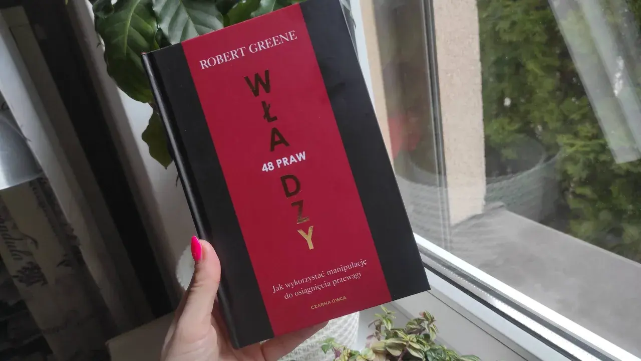 Dłoń z neonowym różowym lakierem trzyma książkę "48 praw władzy" Roberta Greene'a.