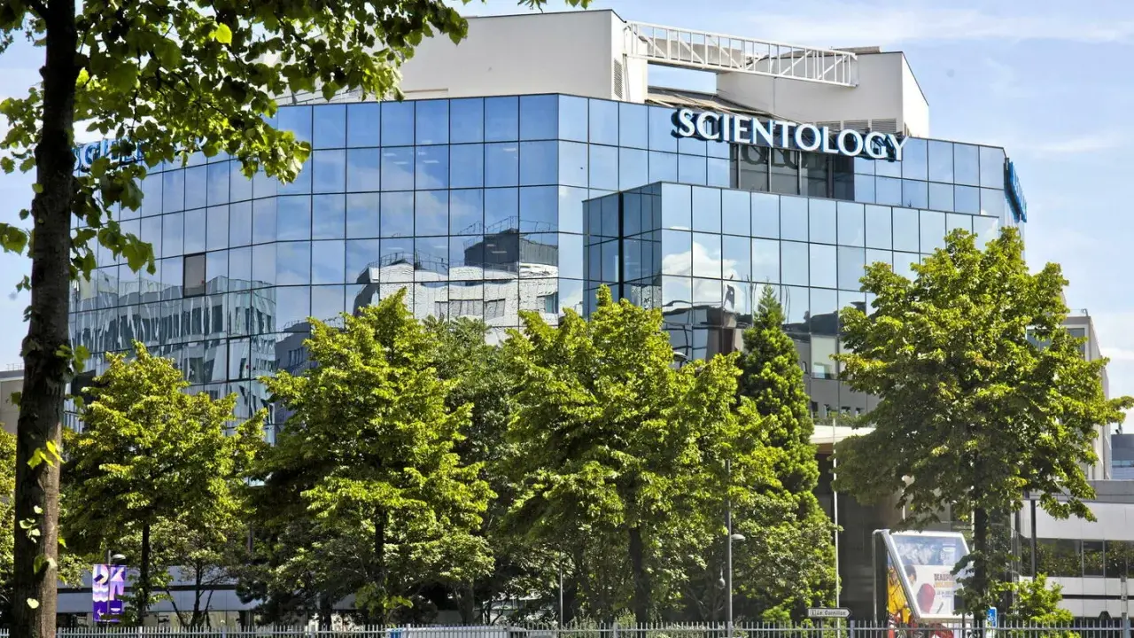 Bâtiment moderne aux façades vitrées reflétant le ciel bleu, avec le nom "SCIENTOLOGY" inscrit en lettres bleues lumineuses. Des arbres verdoyants entourent la structure.