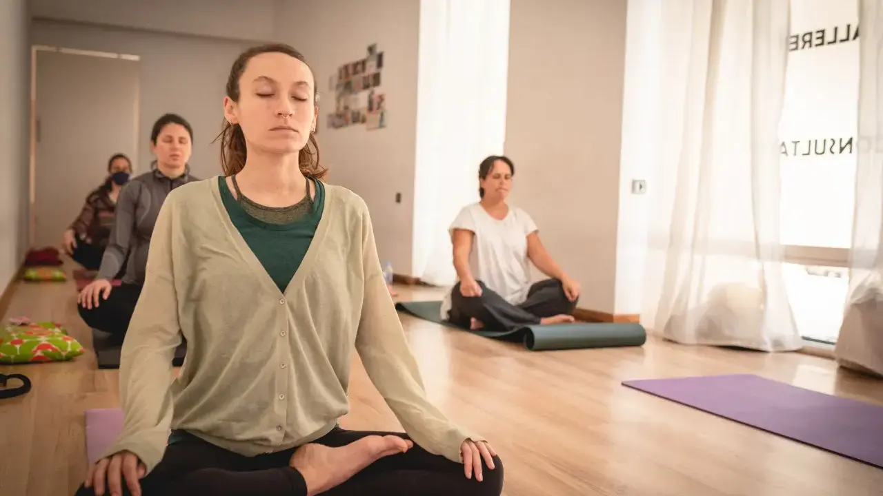Mujeres meditando en clase de yoga. Una de ellas, en primer plano, parece estar en el **primer trimestre de embarazo**, buscando calma y conexión.