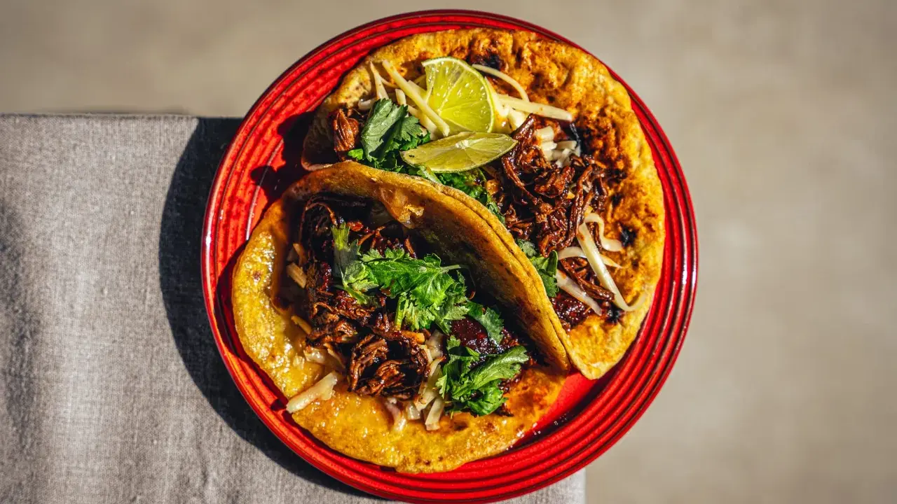 Zwei saftige Birria Tacos auf einem roten Teller, garniert mit Koriander und Limettenspalten.