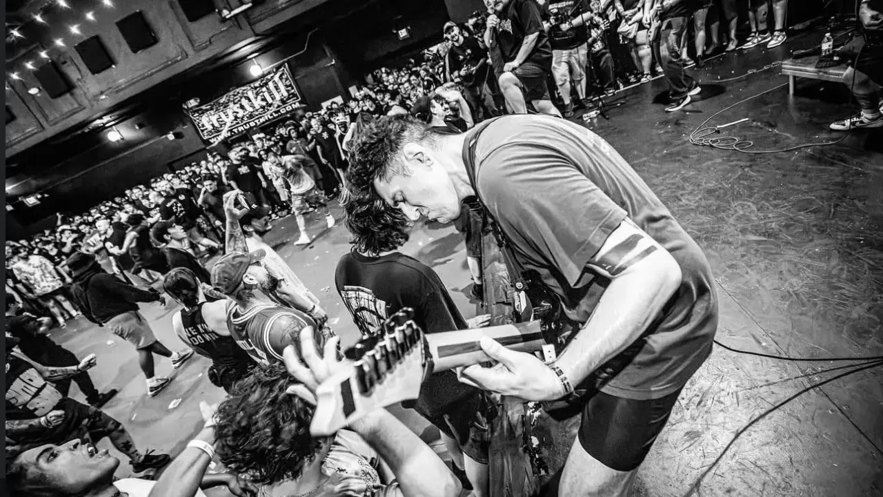 Energetyczny koncert w klubie. Czarno białe zdj. z tłumem szalejącym pod sceną, gdzie gitarzysta pochyla się nad instrumentem.
