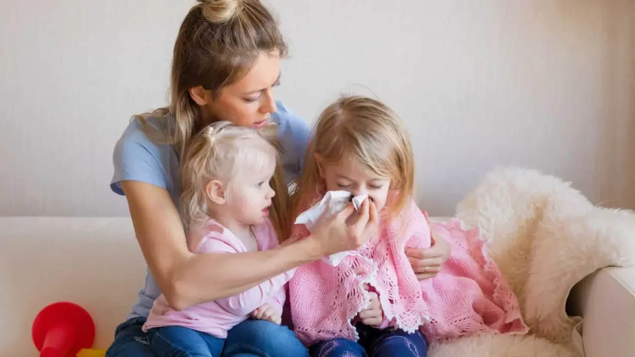 Mama wyciera nos chorej córce, podczas gdy młodsza siostra patrzy. Czy szczepienie mogłoby zapobiec katarowi?