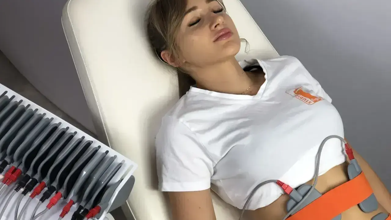 Kobieta leży na kozetce, poddana zabiegowi elektrostymulacji brzucha. Elektrody przymocowane są do jej talii.