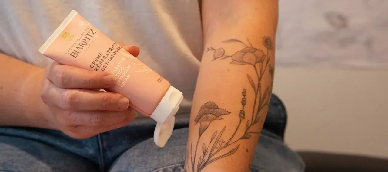 Une main tient un tube de crème réparatrice post-tatouage, prêt à être appliqué sur un bras orné d'un tatouage floral délicat.