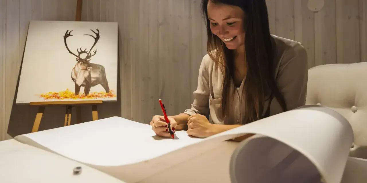 En kvinna ler medan hon ritar på ett stort papper. I bakgrunden syns en oljemålning av en ren. Hon deltar i en oljemålning kurs.