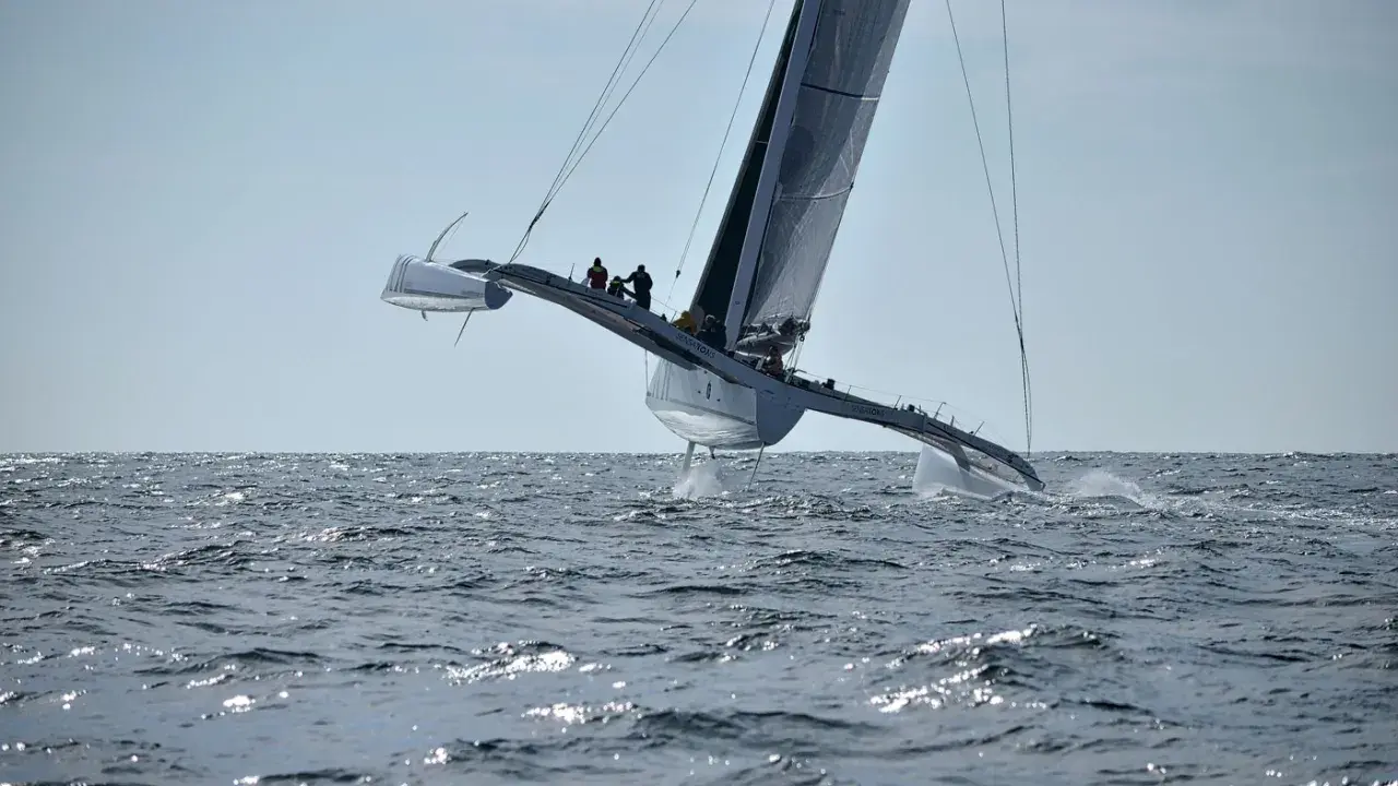 Un maxi trimaran fend les vagues, ses coques latérales hors de l'eau, propulsé par une voile immense. L'équipage est visible sur le pont.