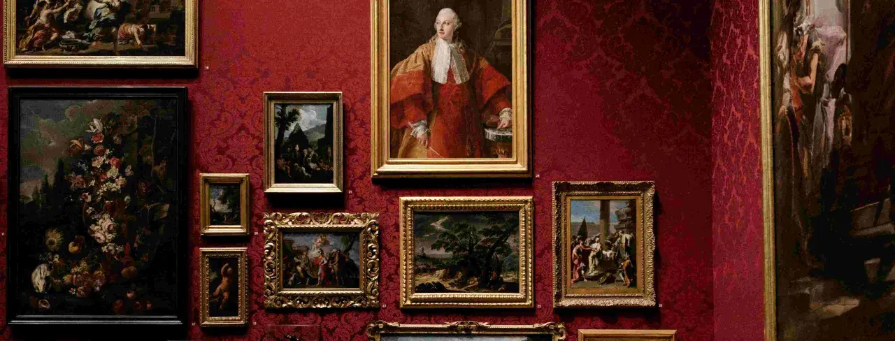Obrazy w bogatym, malarskim stylu zdobią czerwoną, wzorzystą ścianę. Portret mężczyzny w stroju z epoki, bukiet kwiatów i sceny rodzajowe tworzą galerię.