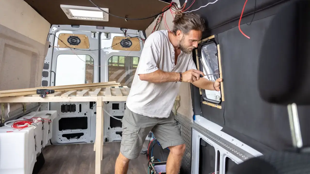 Mężczyzna buduje kamper z osobówki, montując okno. Widać drewniane elementy łóżka i instalację elektryczną.