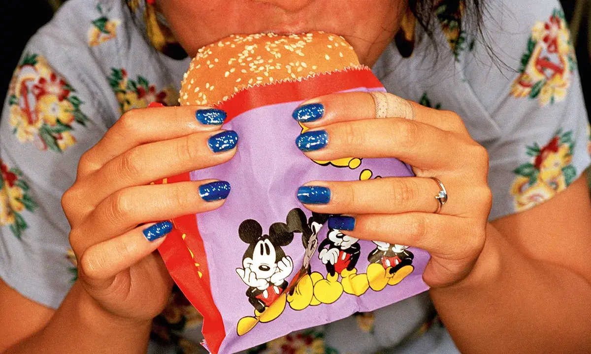 Mani con smalto blu intenso tengono un hamburger in un involucro con Topolino, in uno stile tipico di Martin Parr.