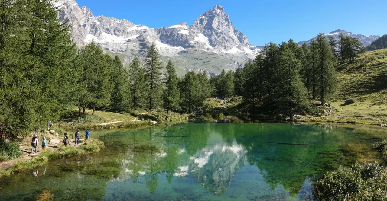 Un gruppo di escursionisti ammira il riflesso del Cervino nel lago blu di Cervinia, circondato da pini e montagne innevate.