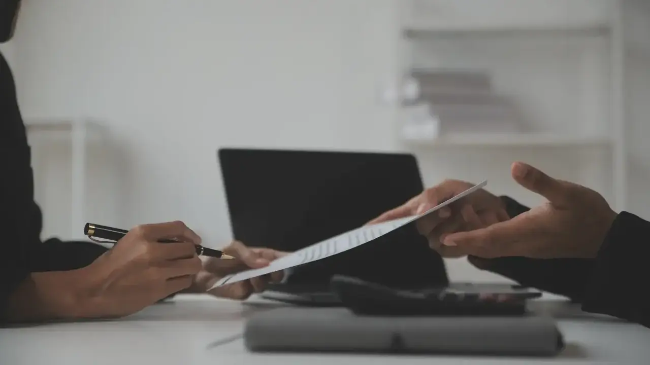 Dwie osoby omawiają dokument, jedna wskazuje piórem na klauzulę CV. Obok leży laptop i kalkulator.
