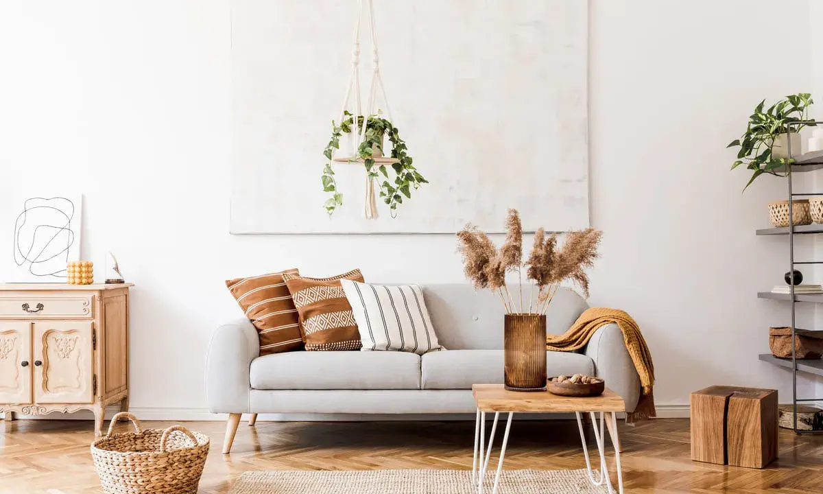 Un salón moderno con un sofá gris, cojines étnicos y una manta mostaza. La decoración se completa con una planta colgante y un jarrón con hierbas secas.