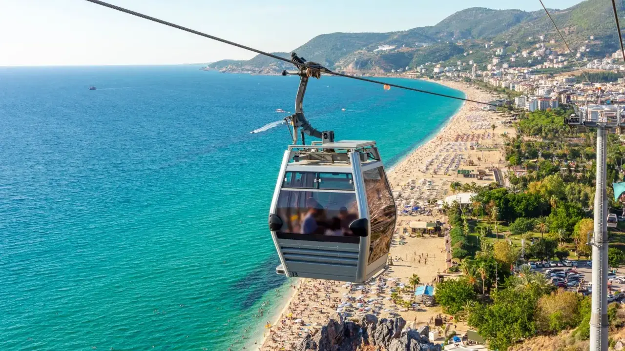 Seilbahn über dem belebten Alanya Strand mit türkisfarbenem Meer und vielen Sonnenanbetern.