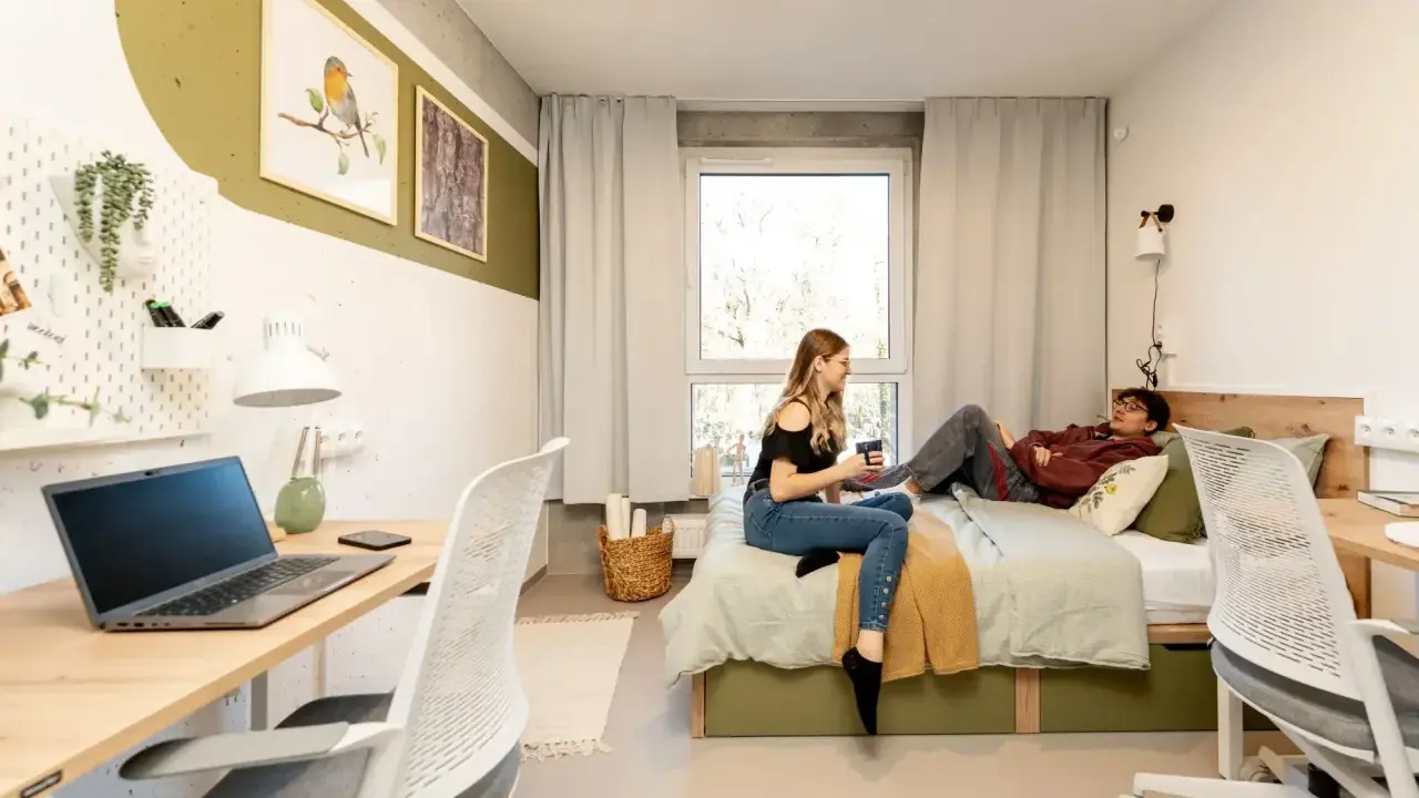 Dwoje studentów w pokoju, marzących o darmowych studiach zaocznych. Na biurku laptop, na łóżku relaks.