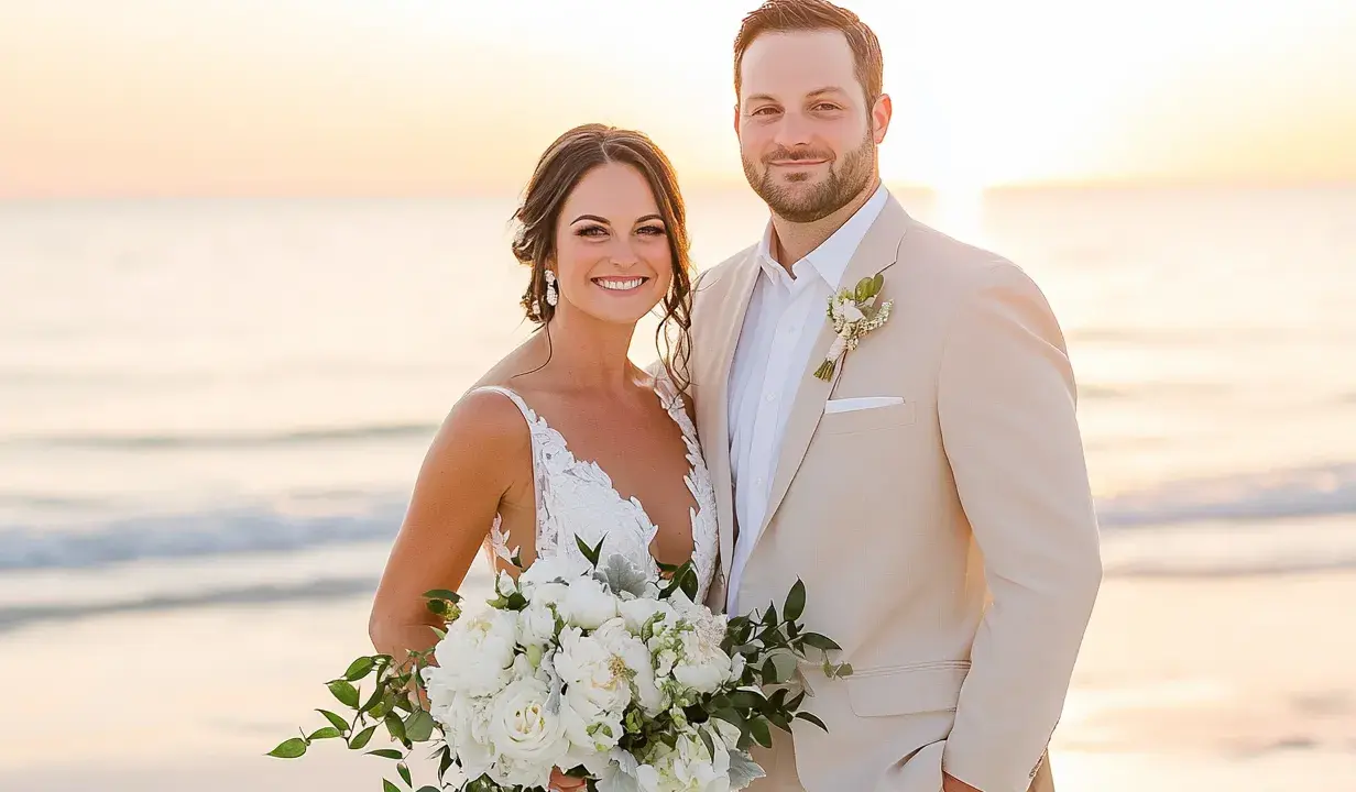 Groom in a tan suit, perfect for what to wear to beach wedding male, smiles with bride holding bouquet at sunset.