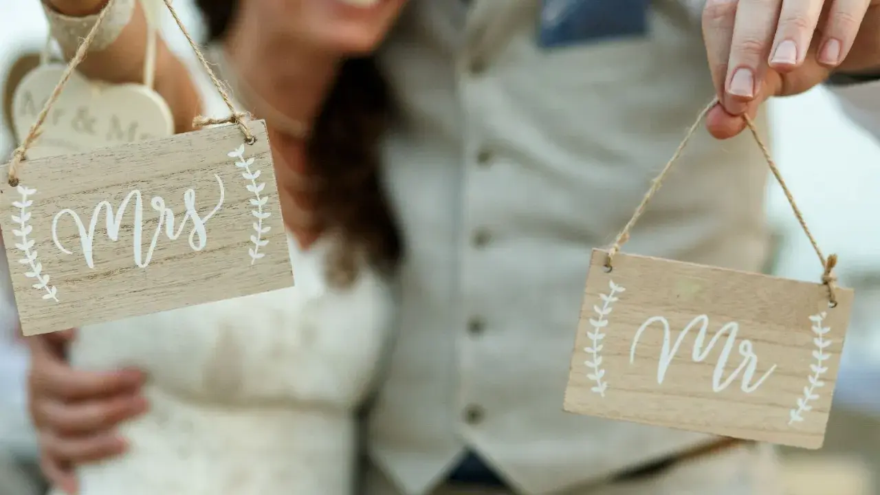 Para Młoda trzyma tabliczki "Mrs" i "Mr", symbolizujące zmianę nazwiska po ślubie.
