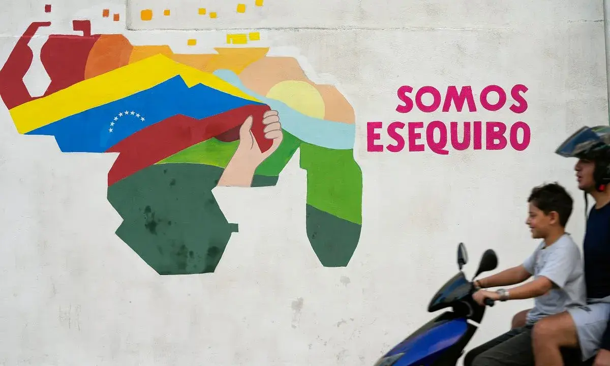 Mural przedstawiający mapę Wenezueli z flagą i hasłem "Somos Esequibo". Dwóch chłopców na skuterze przejeżdża obok.