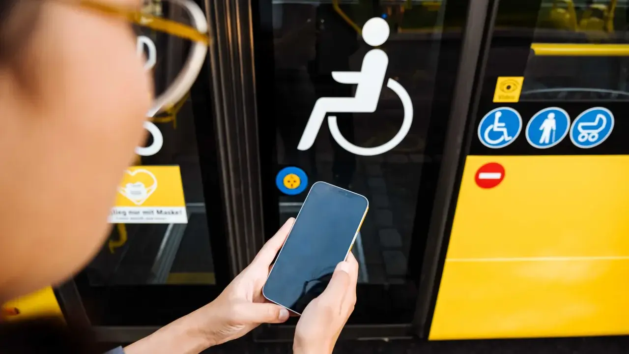 Osoba trzyma telefon przed drzwiami autobusu z symbolem wózka inwalidzkiego. To ułatwia podróżowanie, gdy masz legitymację osoby niepełnosprawnej.