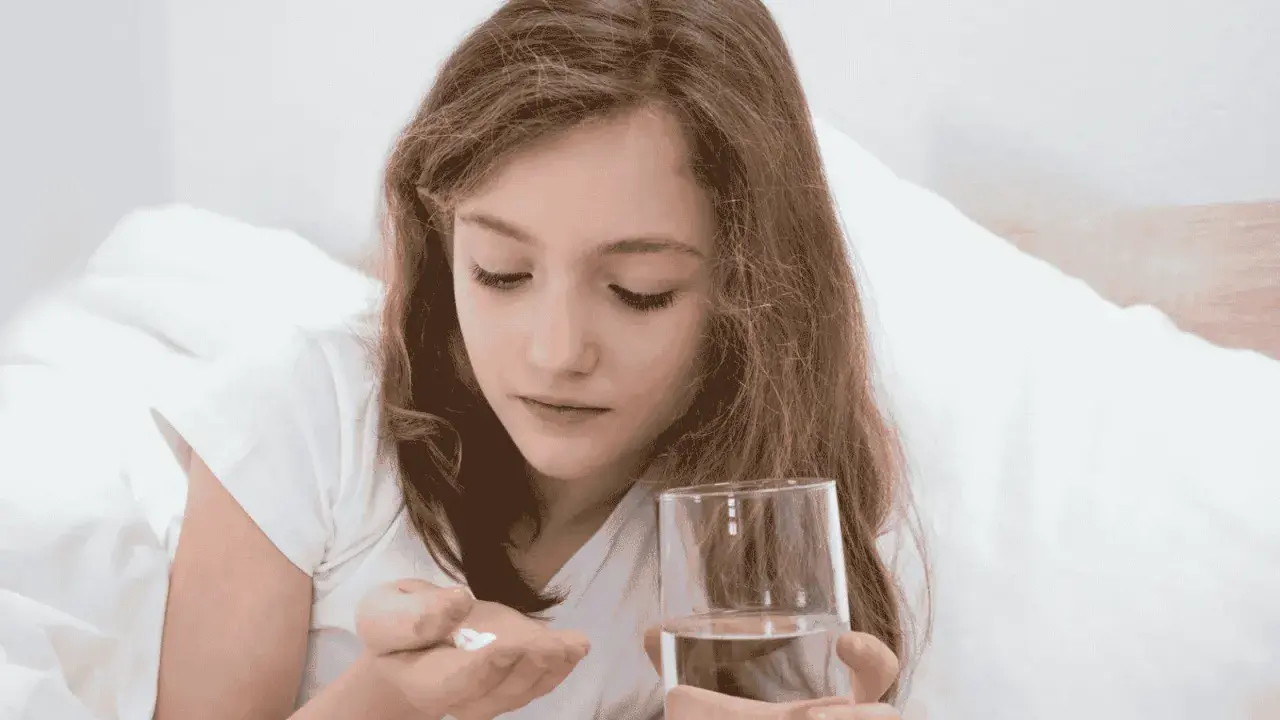 Dziewczynka w łóżku trzyma w dłoni tabletki i szklankę wody. Uważnie patrzy na białe tabletki, które mogą być przeterminowany paracetamol dla dzieci.