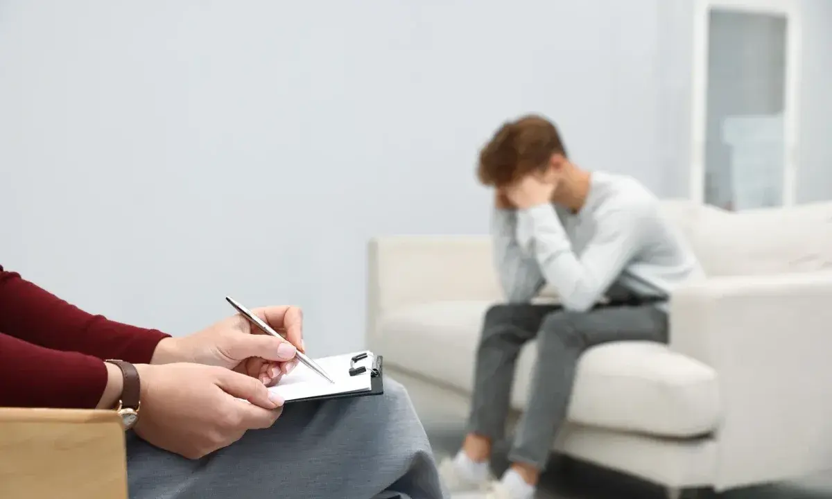 Młody mężczyzna siedzi na kanapie, ukrywając twarz w dłoniach, przeżywając cierpienie psychiczne. Obok niego terapeuta notuje coś na kartce.