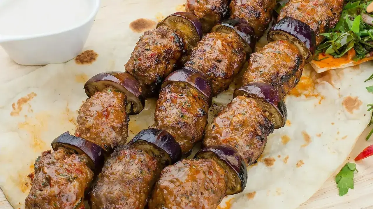 Kofta kebab z bakłażanem na patyku, podany na płaskim chlebie z sosem.