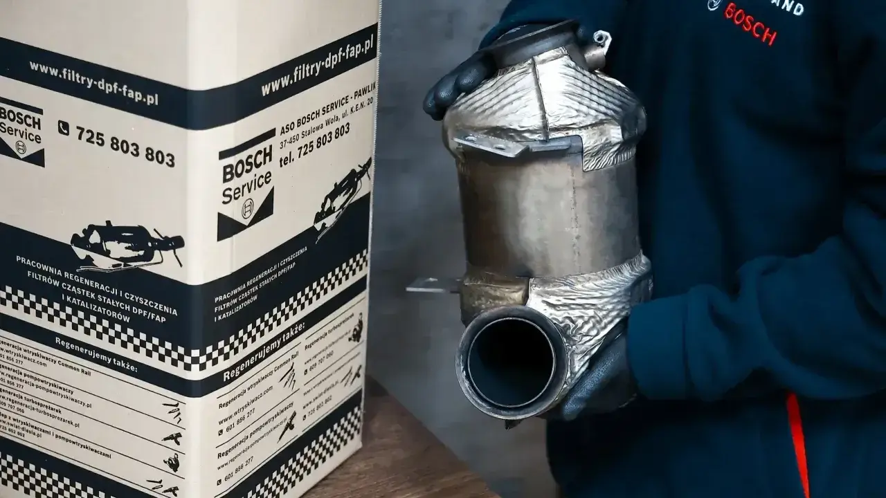 Osoba w rękawiczkach trzyma filtr DPF/FAP. Obok stoi karton z napisem "www.filtry-dpf-fap.pl", sugerującym, że to miejsce zajmuje się regeneracją.