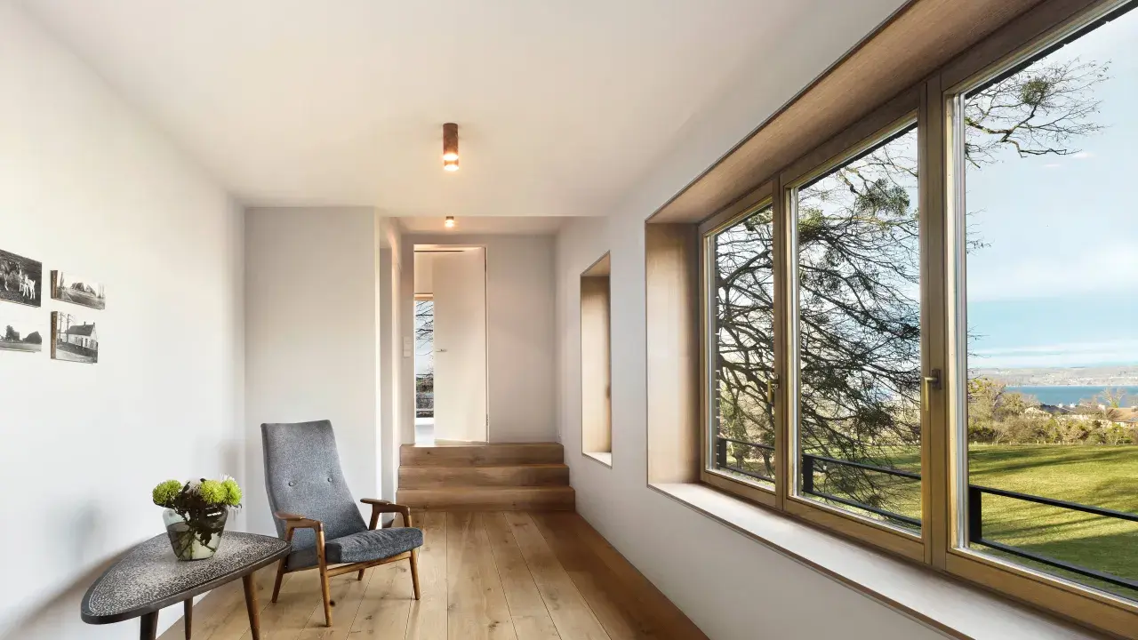 Interior moderno con suelo de madera, sillón gris y gran ventanal. El marco de madera de la ventana, que parece recién restaurado, enmarca un paisaje exterior.