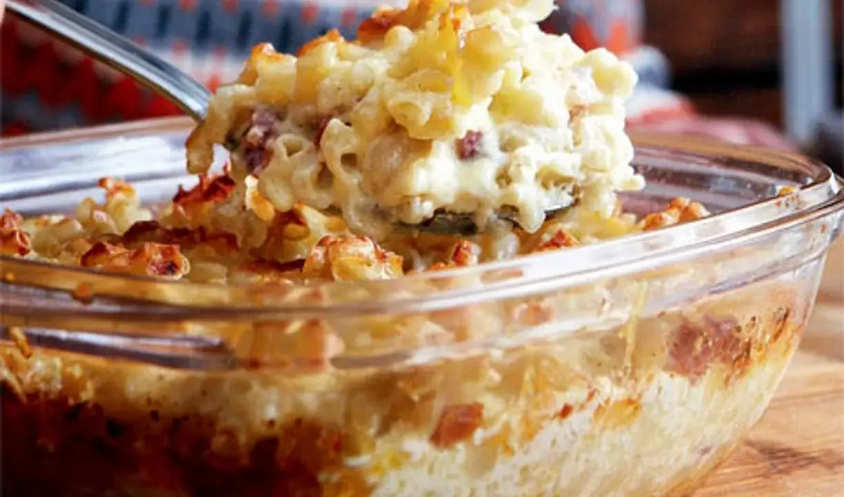 En sked lyfter upp en portion krämig, gammaldags makaronipudding ur en glasform.