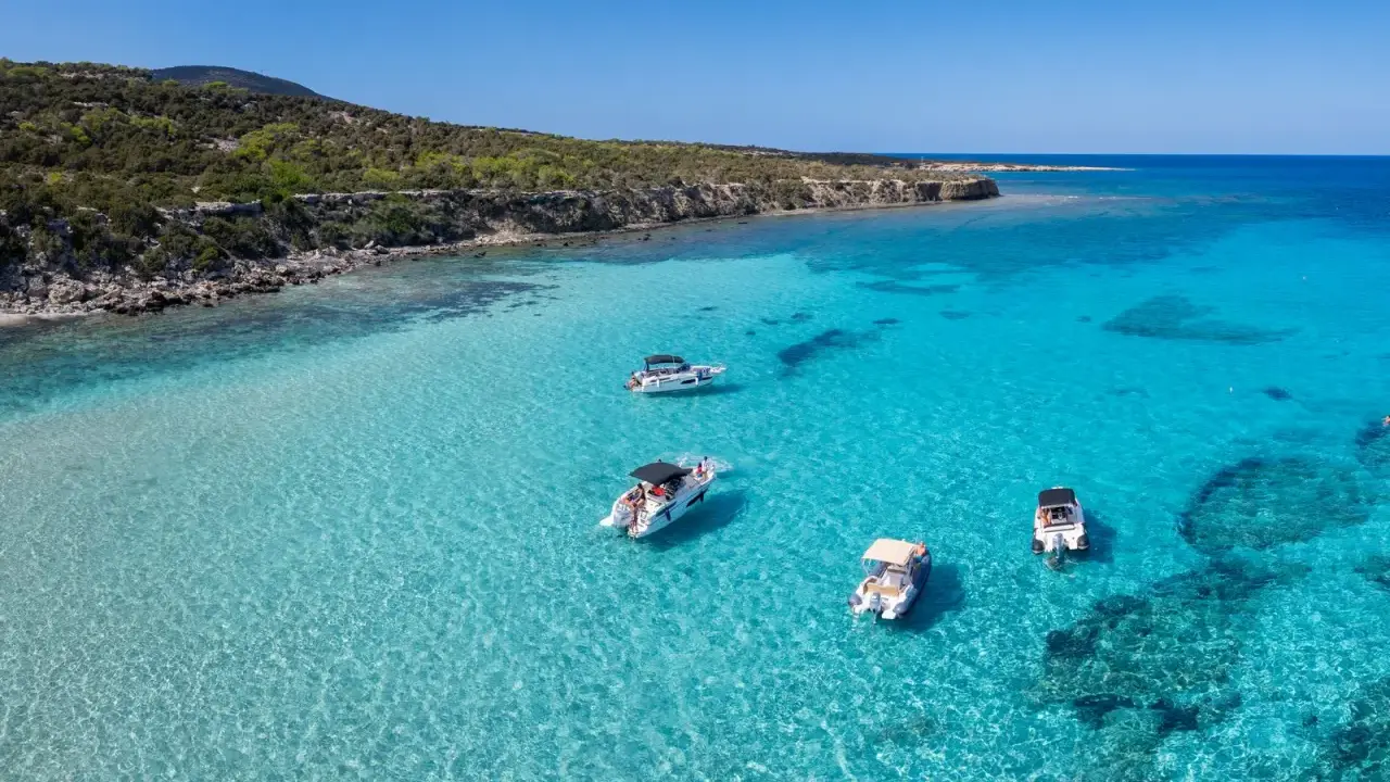 Traumhafter zypern strand mit kristallklarem Wasser und vier Booten, die vor der felsigen Küste ankern.