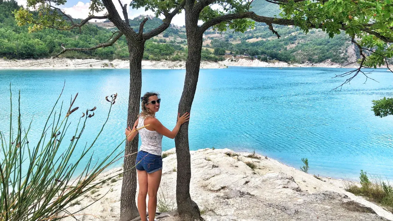 Donna sorridente abbraccia un albero con un lago turchese sullo sfondo. Una perfetta gita fuori porta nelle Marche.