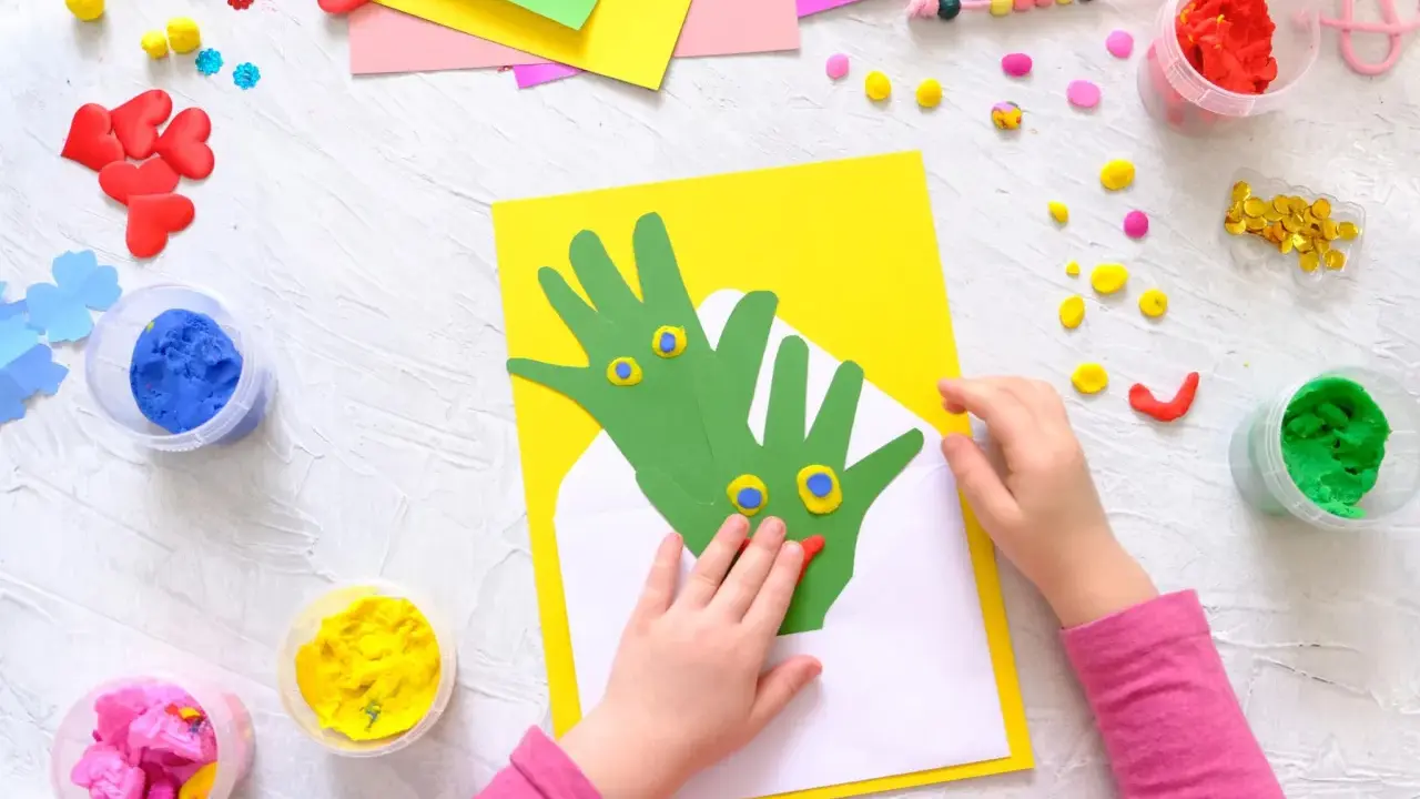 Kreatives Basteln mit Kleinkindern: Hände werden zu grünen Drachen auf gelbem Papier, umgeben von bunter Knete und Bastelmaterialien.