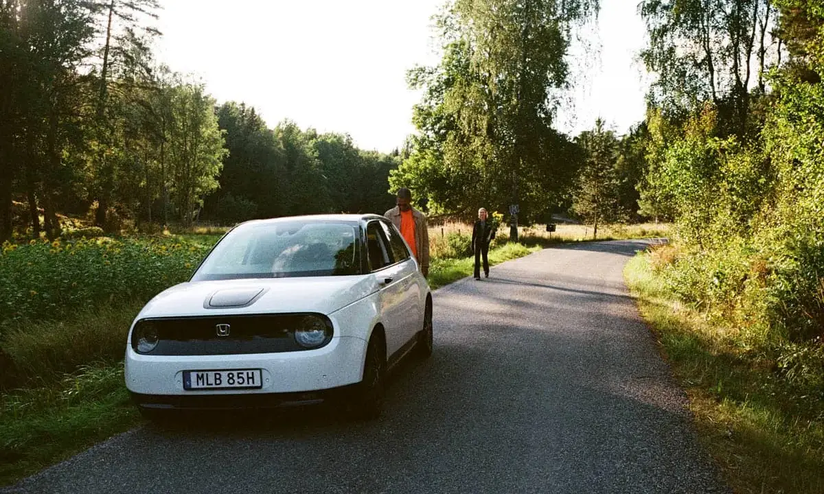 Vit elbil parkerad vid en skogsväg, två personer står vid sidan. En perfekt start på en roadtrip Sverige.