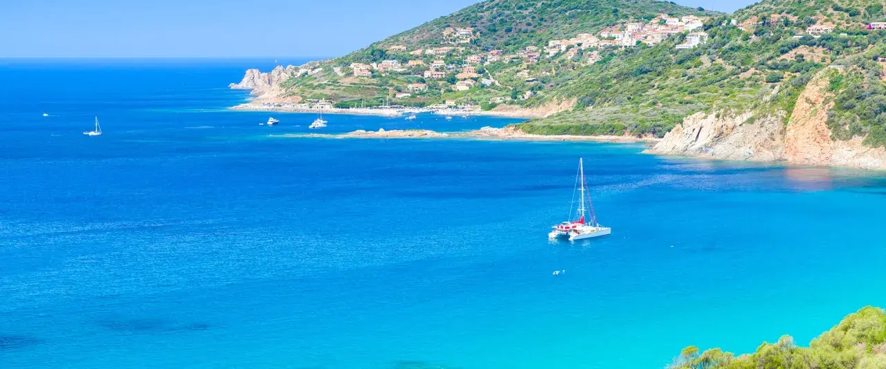Un catamaran blanc avec des accents rouges navigue sur des eaux bleues cristallines, près d'une côte verdoyante et d'un village pittoresque.
