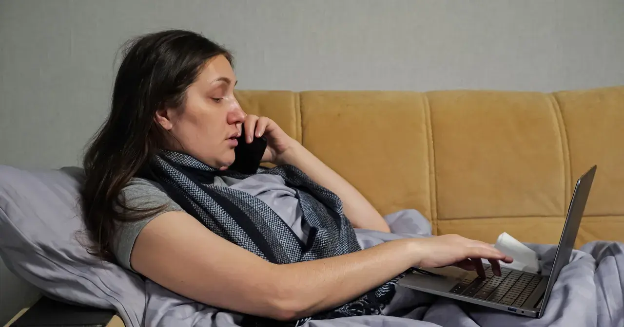 Kobieta w łóżku, rozmawiająca przez telefon i pracująca na laptopie, zastanawia się, na ile dni opłaca się brać zwolnienie lekarskie.