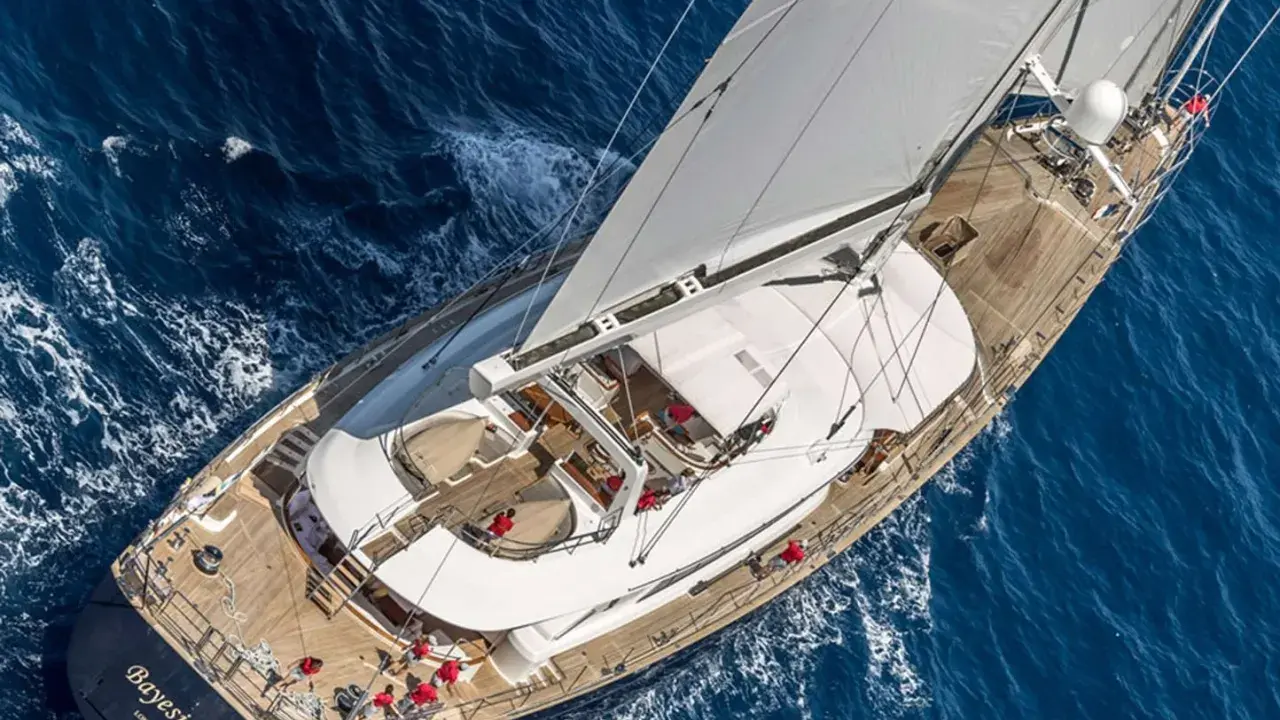 Le yacht "Bayesian" navigue sur des eaux bleues, ses voiles blanches gonflées par le vent. L'équipage en rouge s'affaire sur le pont en bois.