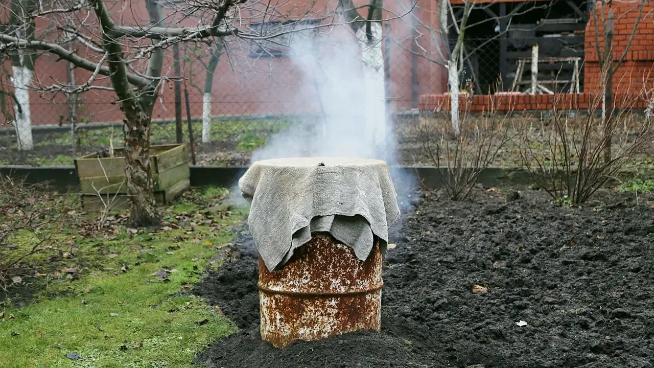 Dym unosi się z beczki, która służy jako prowizoryczna wędzarnia. Wokół niej widać ziemię i trawę.